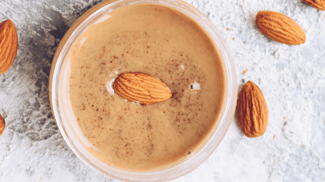 Creamy almond butter in a glass jar with whole almonds on a textured surface – a nutritious, protein-rich spread packed with healthy fats and antioxidants.