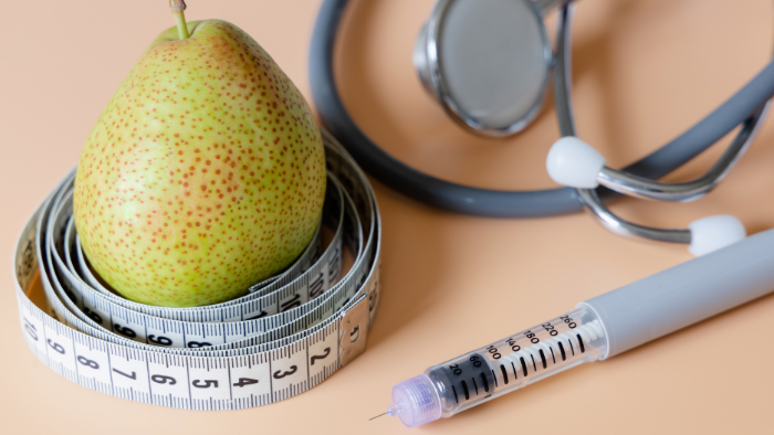 A pear wrapped in a measuring tape, a stethoscope, and a syringe on a beige background, symbolizing weight management and medical treatment.